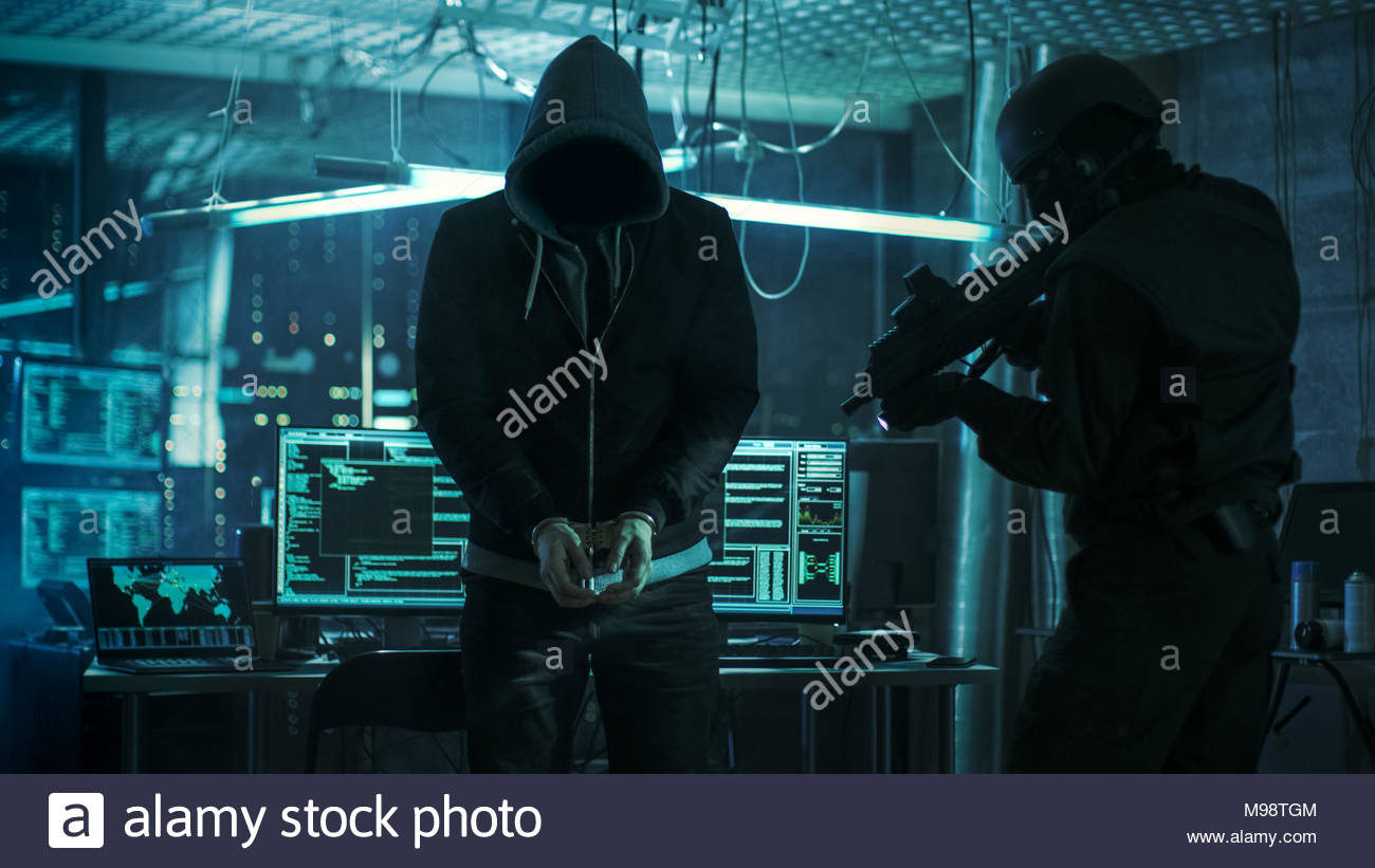 Handcuffed-masked-hacker-is-standing-and-guarded-by-fully-armed-special-forces-soldier-theyre-in-hackers-hideout-basement-with-multiple-displays-M98TGM-1